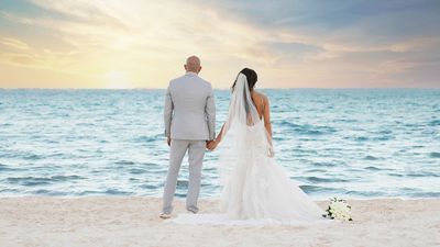 A bride and groom stand hand in hand on a sandy beach, gazing at a serene ocean under a vibrant sunset sky.