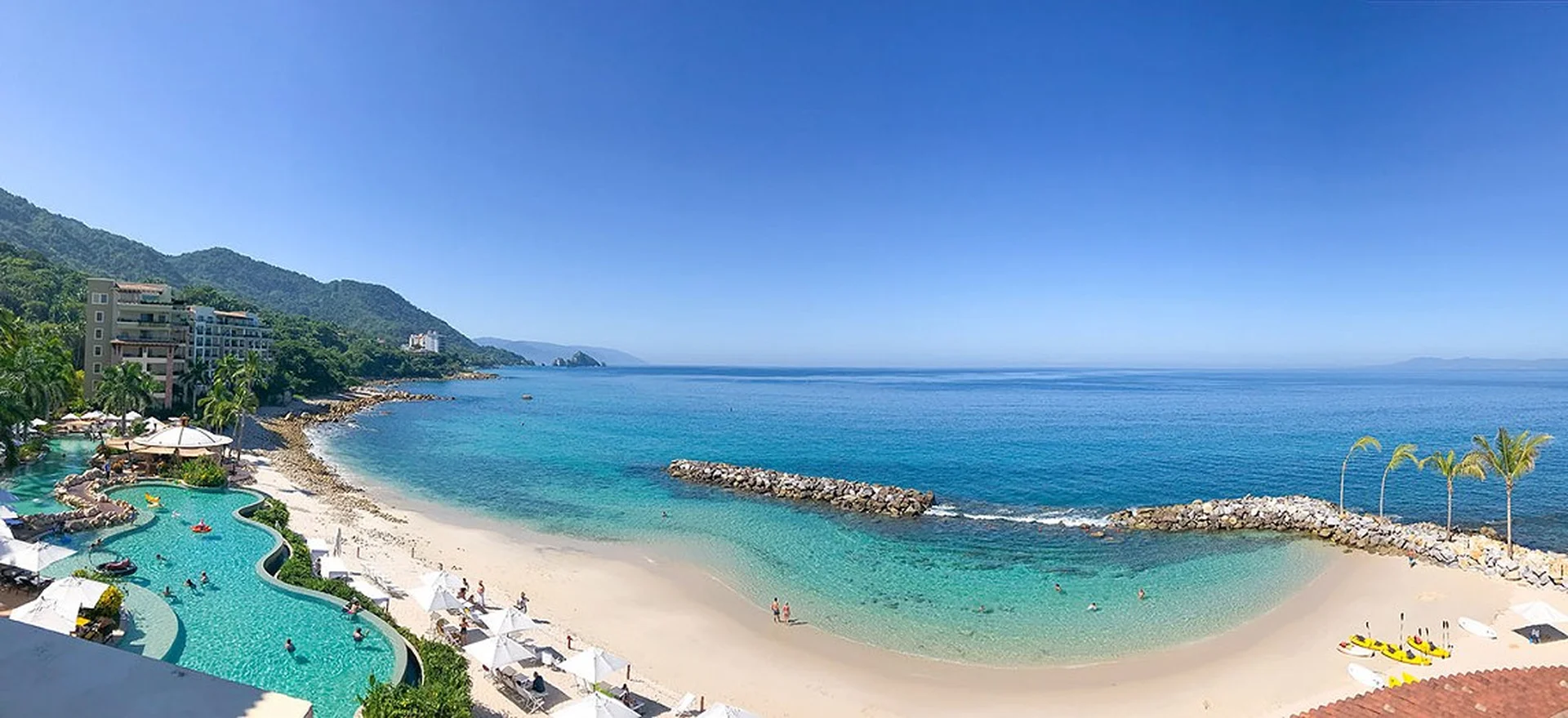 Panoramic view of a tropical beach with clear blue water, a sandy shore, a resort pool, and lush green hills under a clear blue sky.
