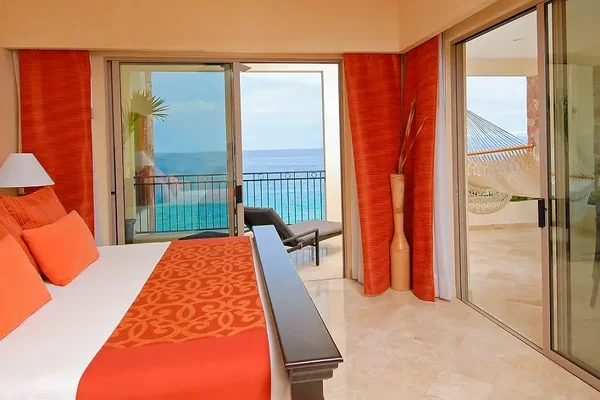 Bright hotel room with an ocean view, featuring a bed with orange accents, sliding glass doors, and a balcony with a hammock and lounge chair.