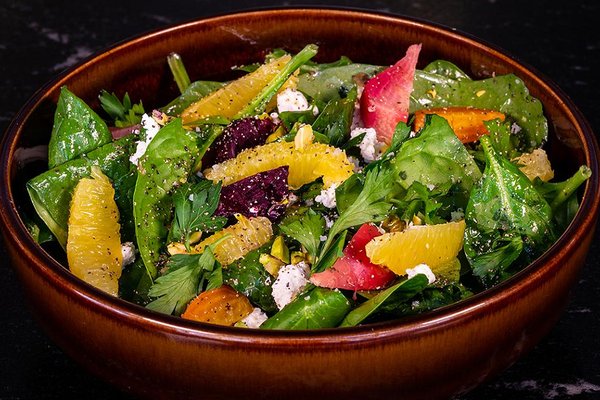 A vibrant salad with spinach, citrus slices, beets, feta cheese, and pistachios in a brown bowl on a dark background.