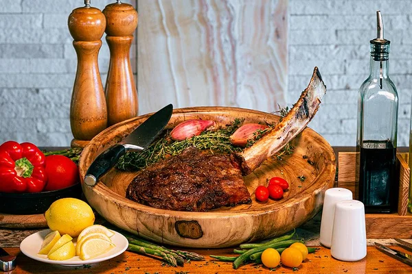 A cooked tomahawk steak on a wooden platter with herbs, cherry tomatoes, and onions, surrounded by peppers, lemons, and condiments.