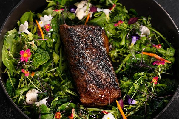 Grilled steak on a bed of mixed greens and vibrant edible flowers in a black pan.