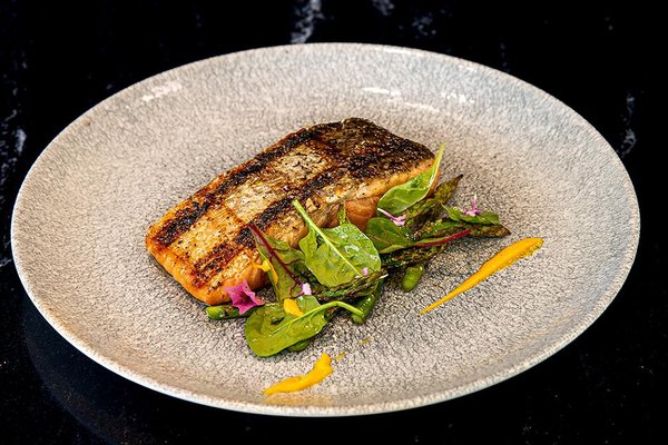 Grilled salmon fillet on a gray plate with mixed greens, asparagus, and yellow sauce garnish on a dark background.
