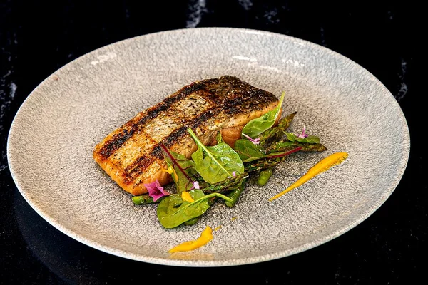 Grilled salmon fillet on a gray plate with mixed greens, asparagus, and yellow sauce garnish on a dark background.