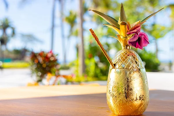 Golden pineapple-shaped cup with a straw and flower garnish on a wooden table, set against a blurred tropical background.