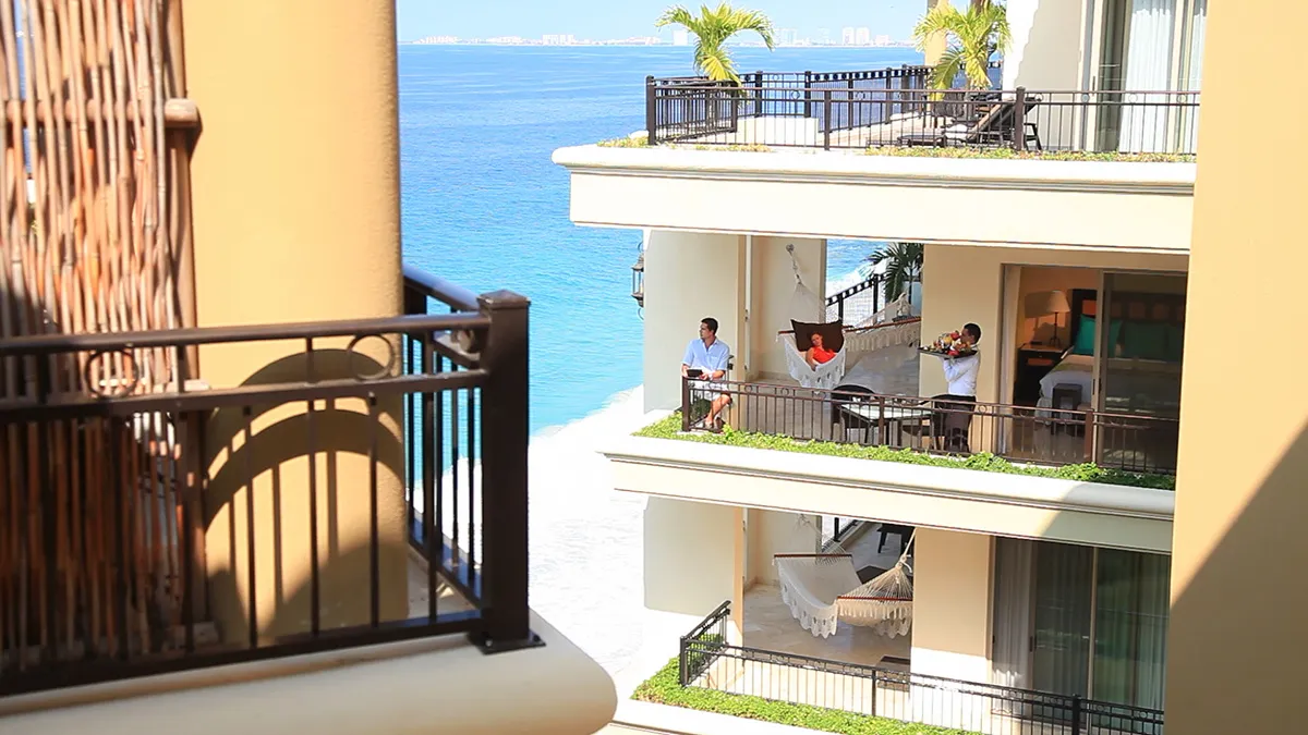 Resort staff preparing a hammock on a luxury balcony overlooking the turquoise sea, with lush greenery and elegant architecture.
