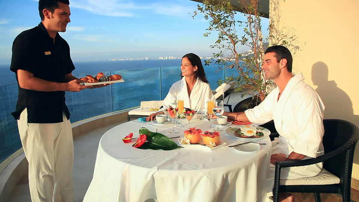 Couple enjoying a luxurious breakfast on a balcony overlooking the ocean, served by a waiter at Garza Blanca Resort, Cancun.