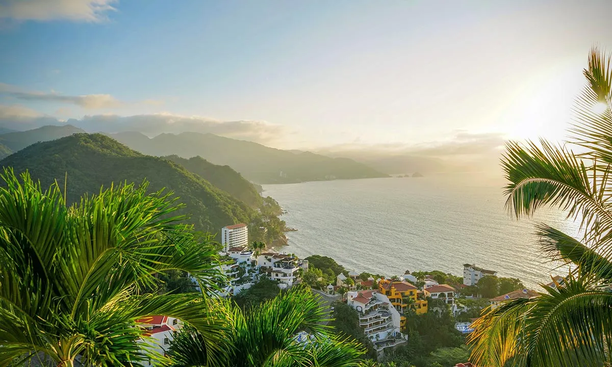 Sunset over lush green hills and serene coastline in Cancun, Mexico, with palm trees framing the picturesque view.