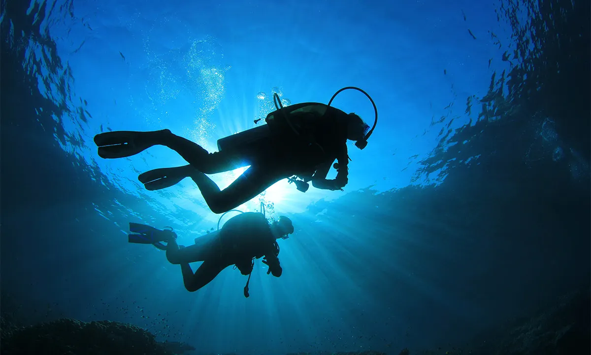 Two scuba divers explore an underwater scene, silhouetted against the sunlit, vibrant blue water.