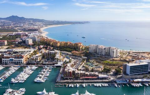 Luxurious Mexican coast: a marina full of yachts, upscale resort buildings along the water, and a long sunlit beach meeting turquoise sea.