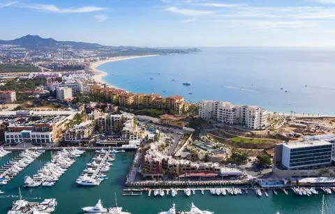 Luxurious Mexican coast: a marina full of yachts, upscale resort buildings along the water, and a long sunlit beach meeting turquoise sea.