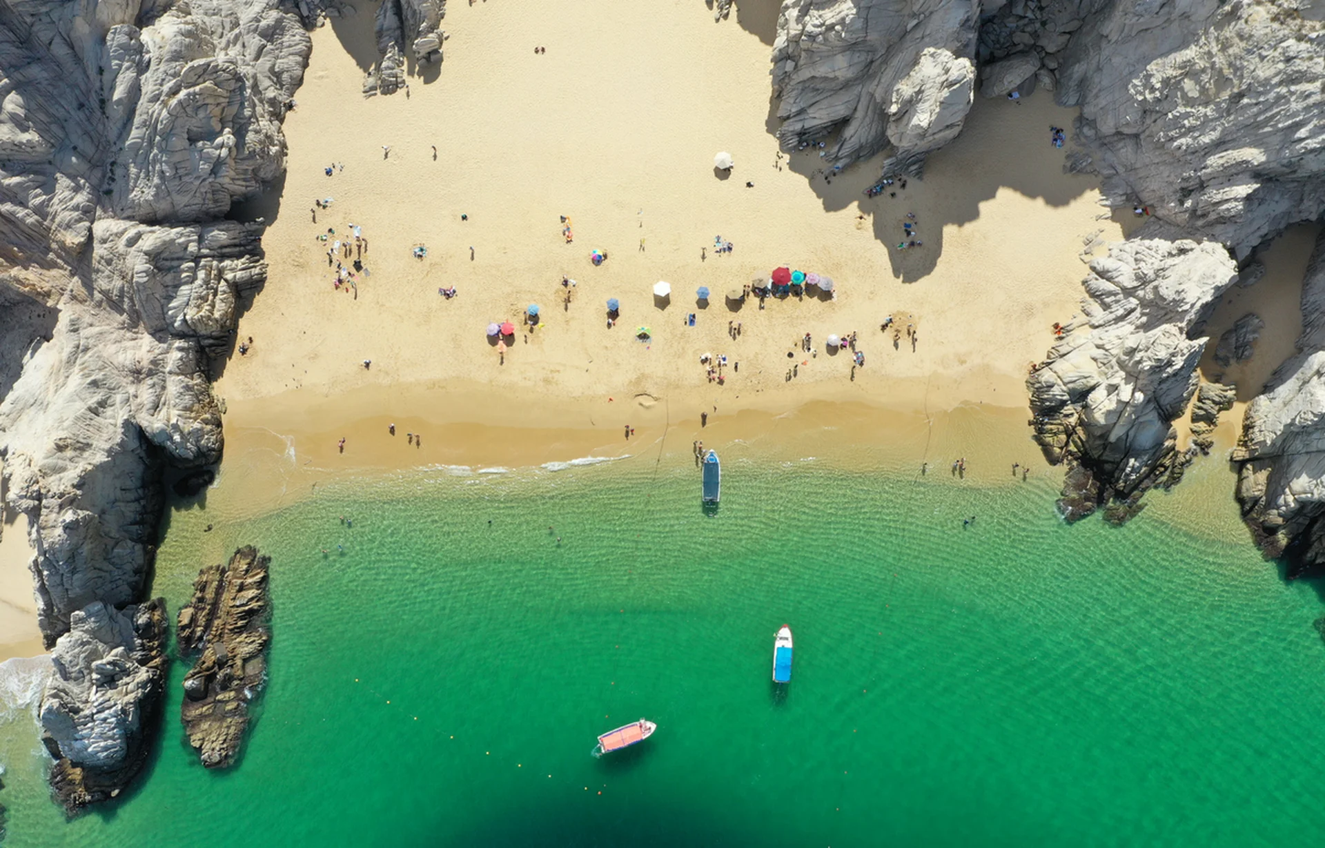 A secluded Mexican cove: golden sand, rugged cliffs, emerald water, anchored boats, and sunbathers under colorful umbrellas at a luxury beach setting.