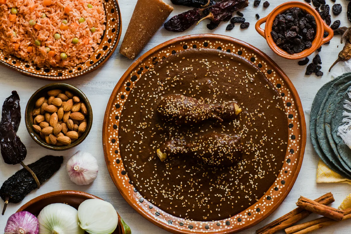 Traditional Mexican mole dish with chicken, surrounded by rice, almonds, garlic, onions, dried chiles, and cinnamon on a rustic table.