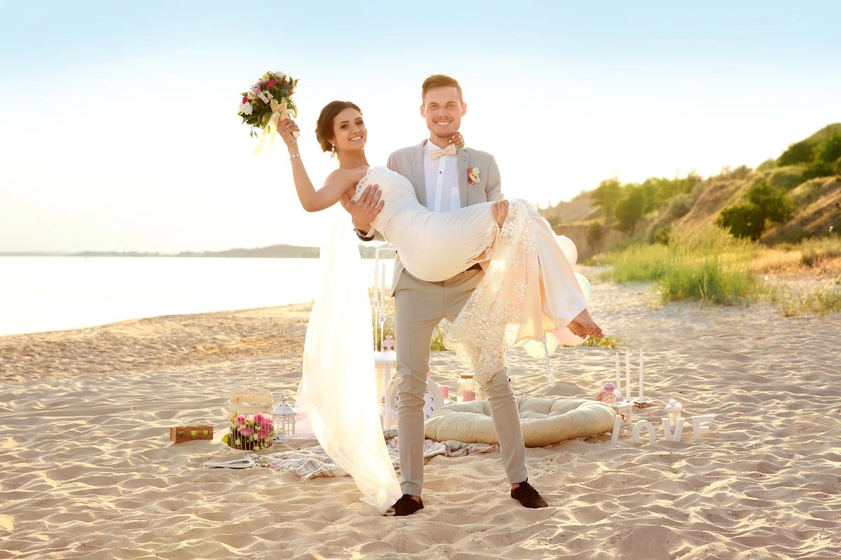 A joyful bride and groom celebrate their beach wedding at sunset, with a picnic arrangement and scenic shoreline in the background.