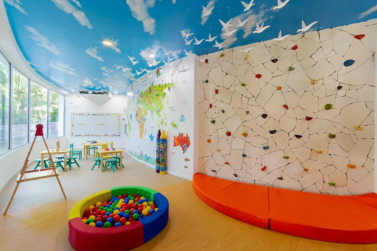 Colorful children's playroom with a climbing wall, ball pit, small tables, and artwork on walls. Bright blue ceiling with white bird decor.