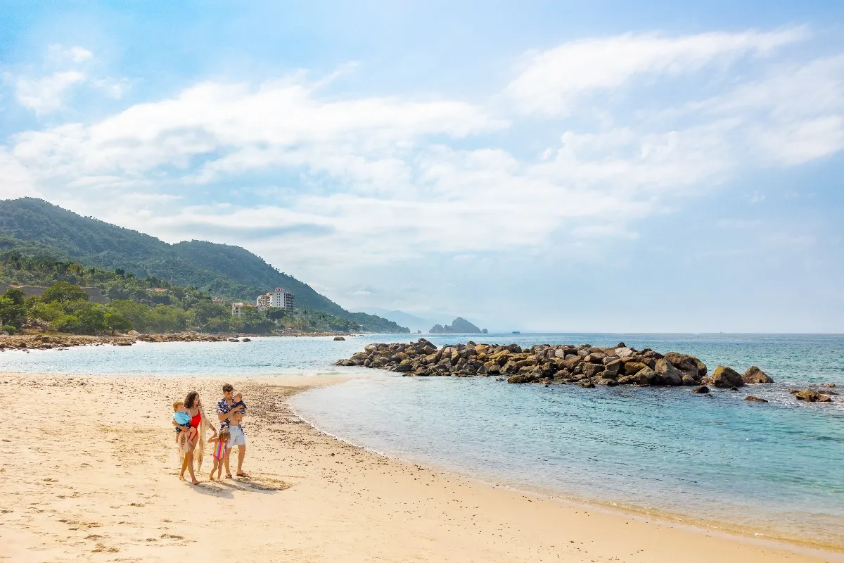 A family walks along a pristine beach with clear blue waters and lush green hills in the background under a partly cloudy sky.
