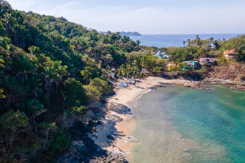 Aerial view of a secluded beach with palm trees and turquoise waters, bordered by lush greenery and small buildings in the distance.