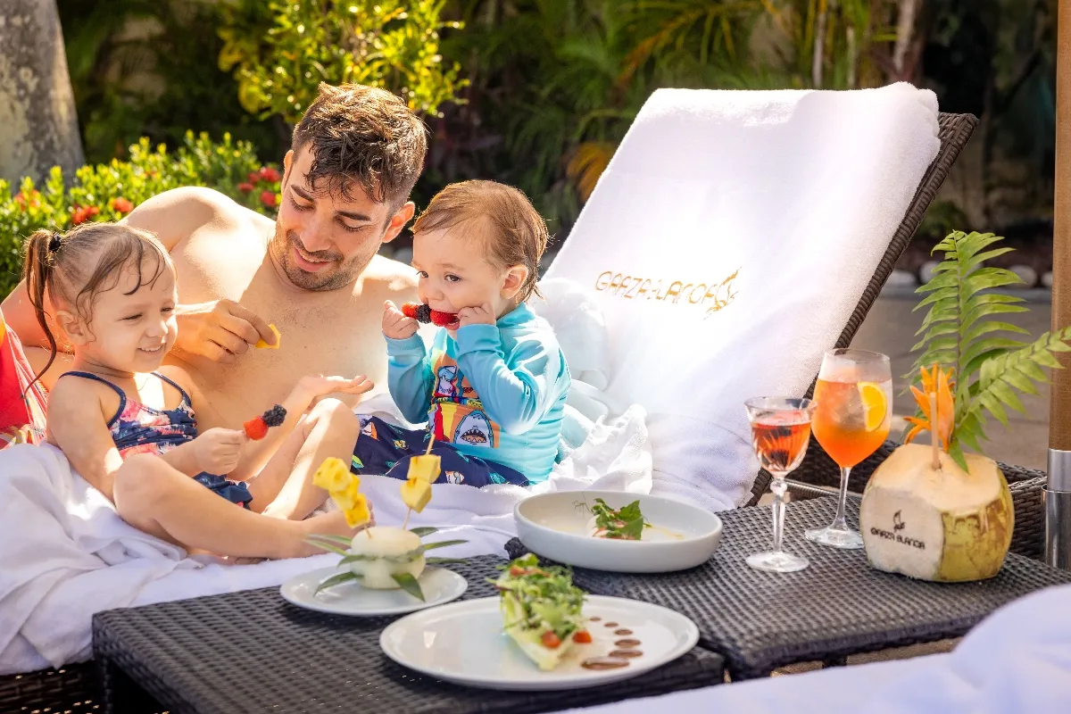 A father and two children enjoy fresh fruit skewers by the pool at Garza Blanca Cancun, with tropical cocktails and vibrant greenery around.