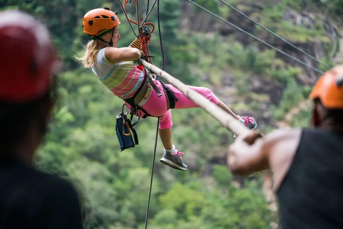 A person enjoying a thrilling zipline ride through lush green forest, surrounded by safety gear and ropes, with others watching.
