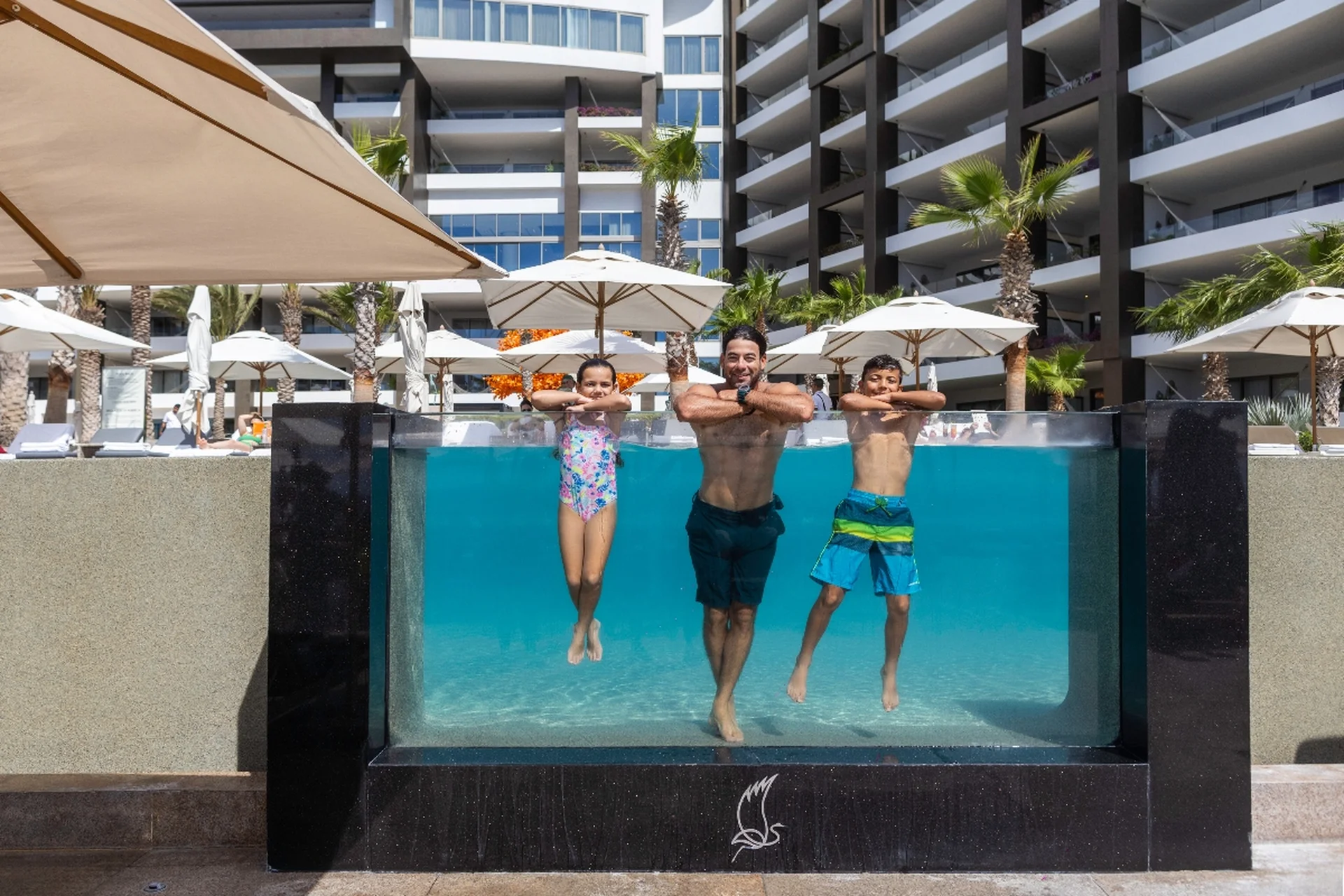 Padre y dos niños flotando en una alberca con paredes de cristal en Garza Blanca Cancún, con sombrillas, palmeras y el moderno resort al fondo.