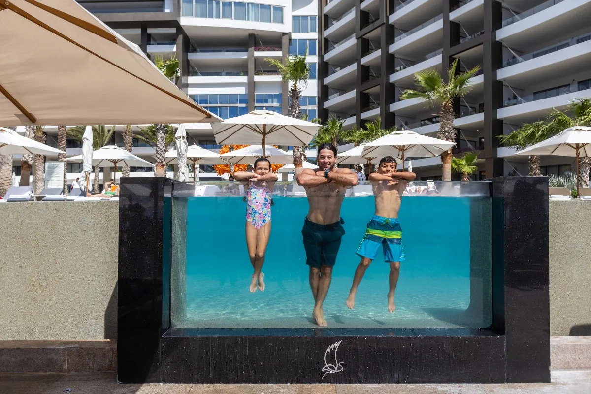 father and two children floating in a glass-walled pool at Garza Blanca Cancun with umbrellas, palms and modern resort behind.