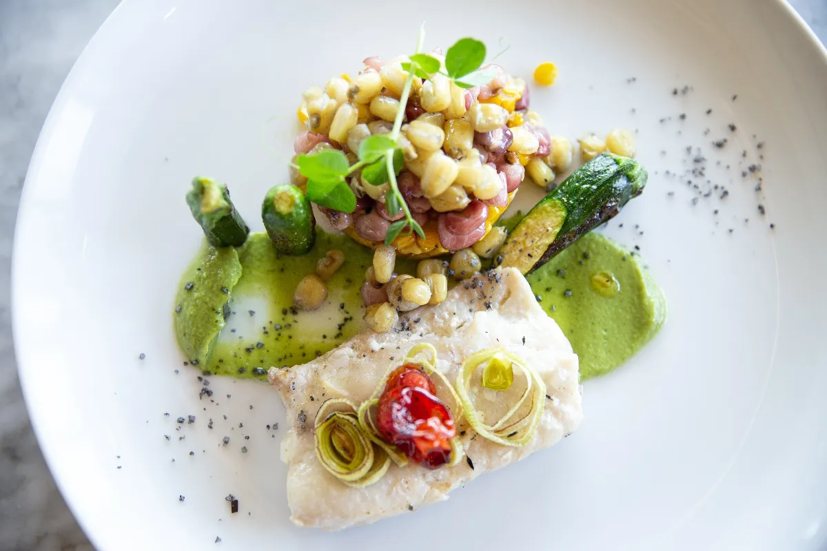 Gourmet dish featuring fish fillet with roasted vegetables, creamy sauce, and corn salad, artfully presented on a white plate.