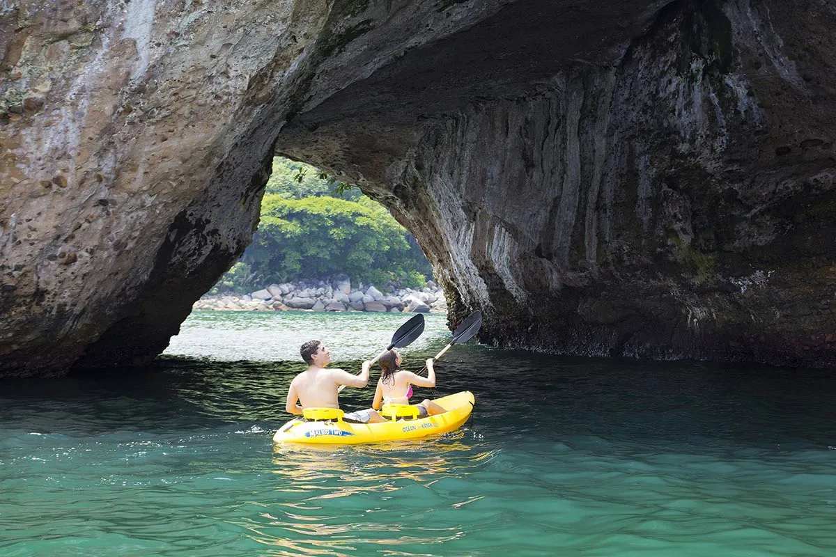 Two people kayaking through a rocky archway over tranquil turquoise waters, surrounded by lush greenery.