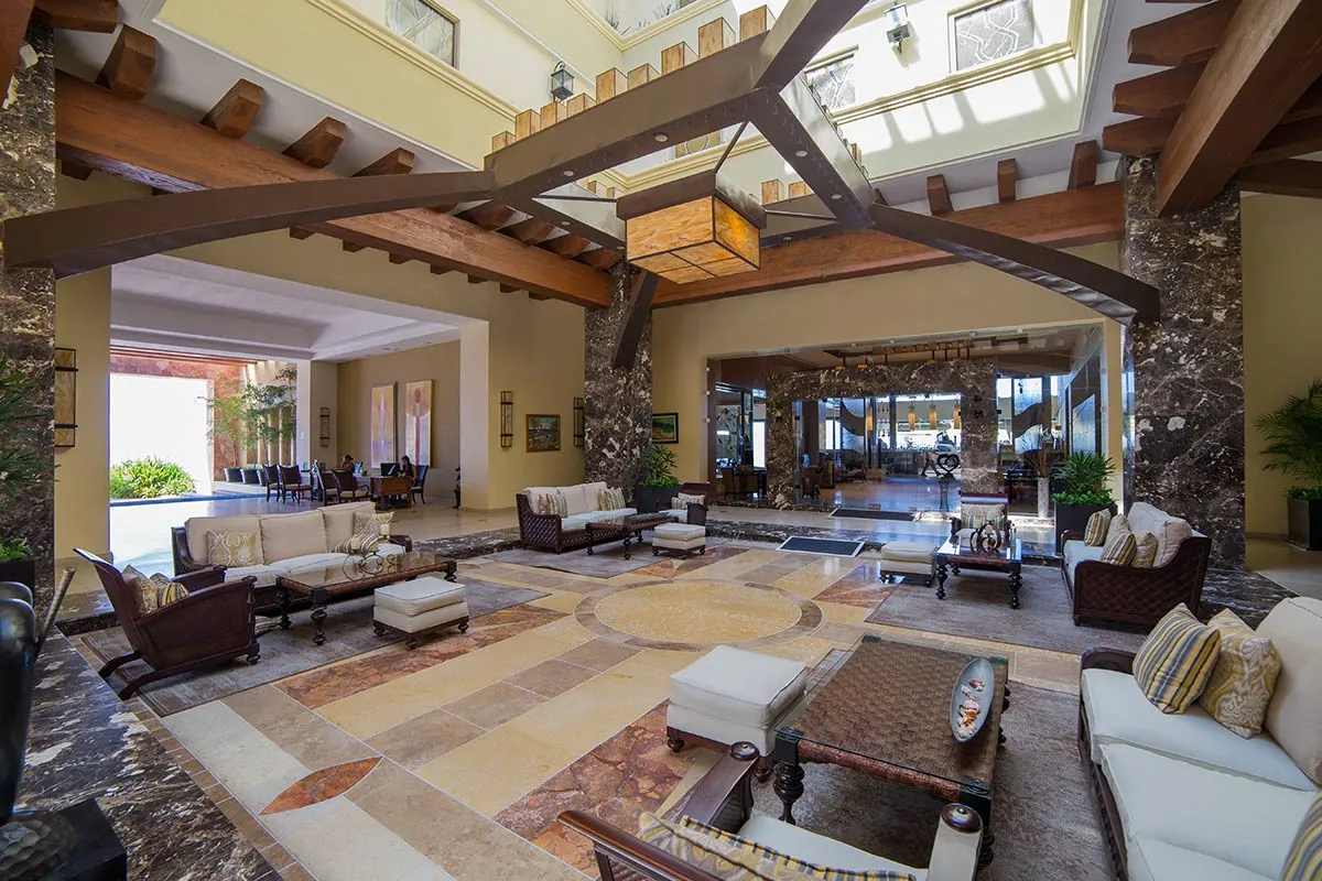 Luxurious resort lobby with elegant wooden beams, plush seating, marble flooring, and a view of the dining area beyond.