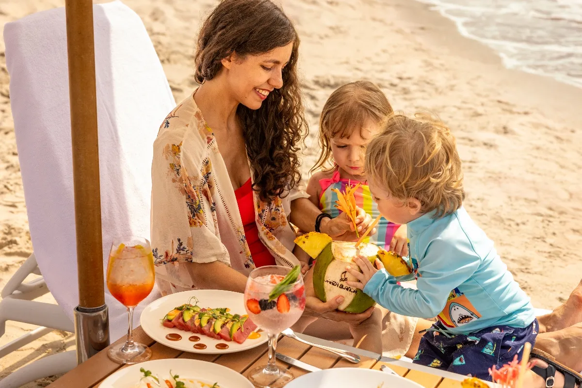 A mother with two children enjoys fresh coconut drinks and gourmet dishes on a sunny beach, surrounded by the serene ocean.