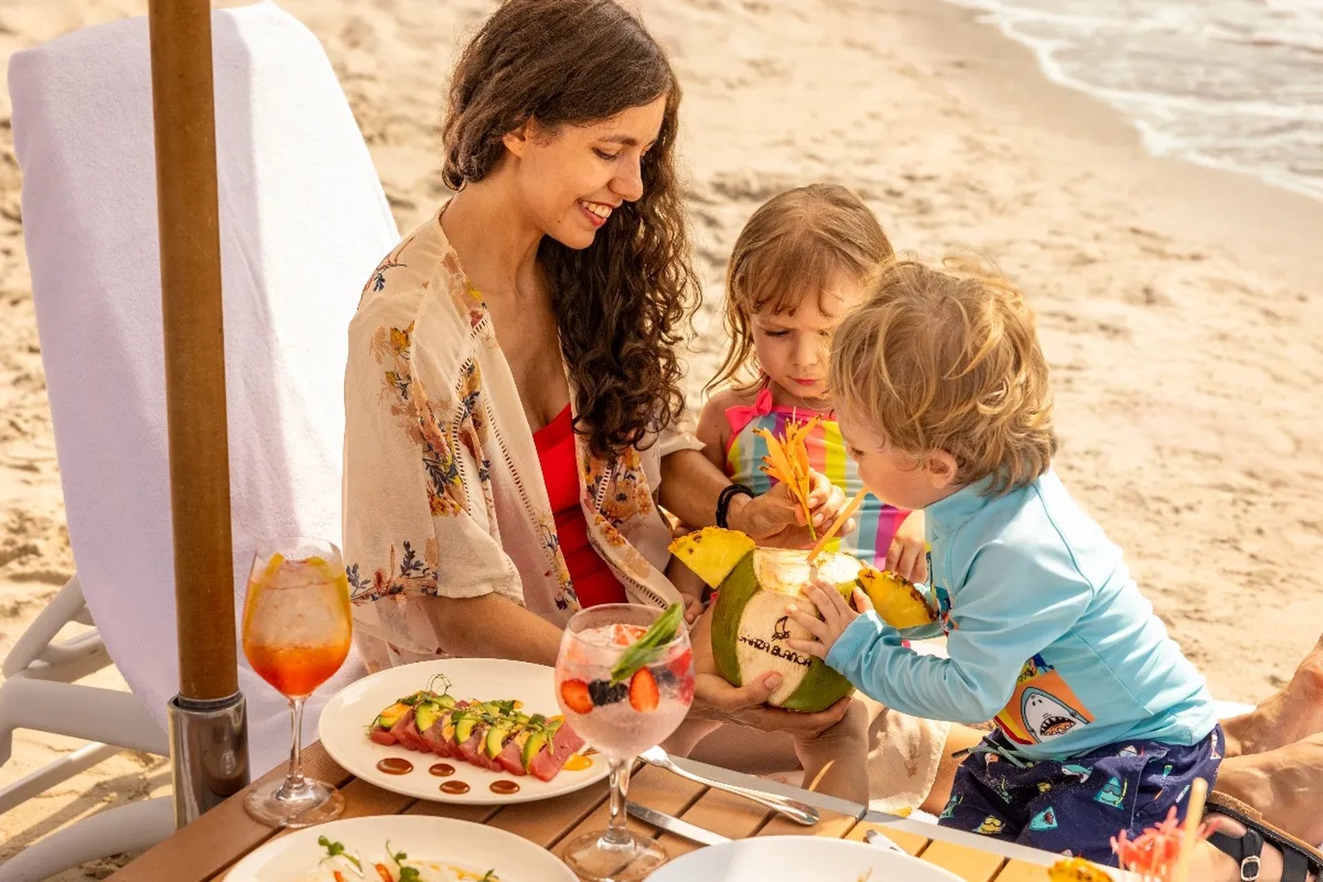 A mother with two children enjoys fresh coconut drinks and gourmet dishes on a sunny beach, surrounded by the serene ocean.