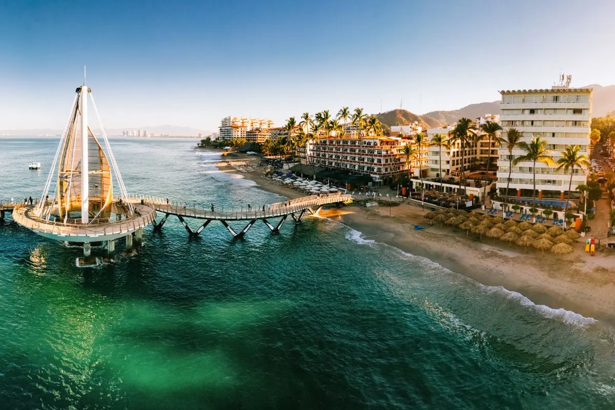 Aerial view of a stunning beachfront with a pier and inviting clear waters, surrounded by vibrant buildings and lush palm trees.
