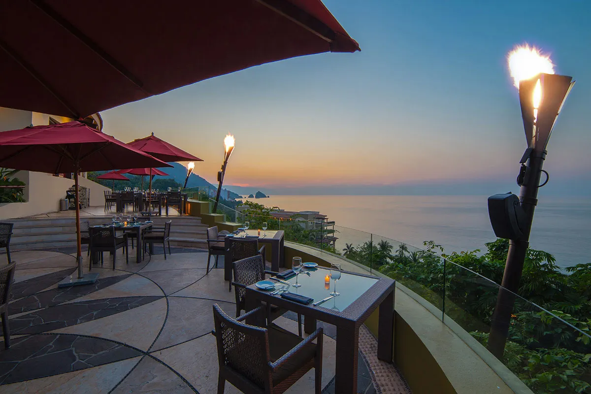 Outdoor dining area at sunset with ocean views, elegant tables, red umbrellas, and tiki torches creating a warm, inviting ambiance.