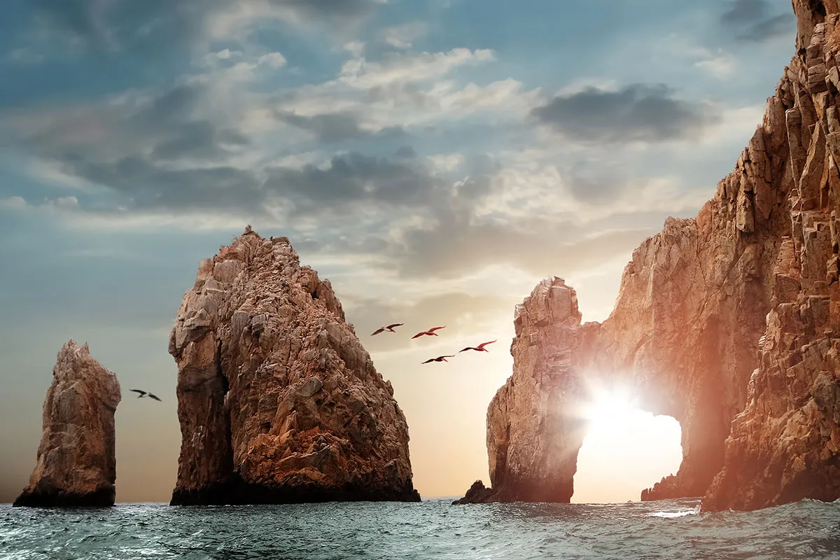Sunset view of El Arco rock formation at Land's End, Cabo San Lucas, with a dramatic sky and birds flying by.