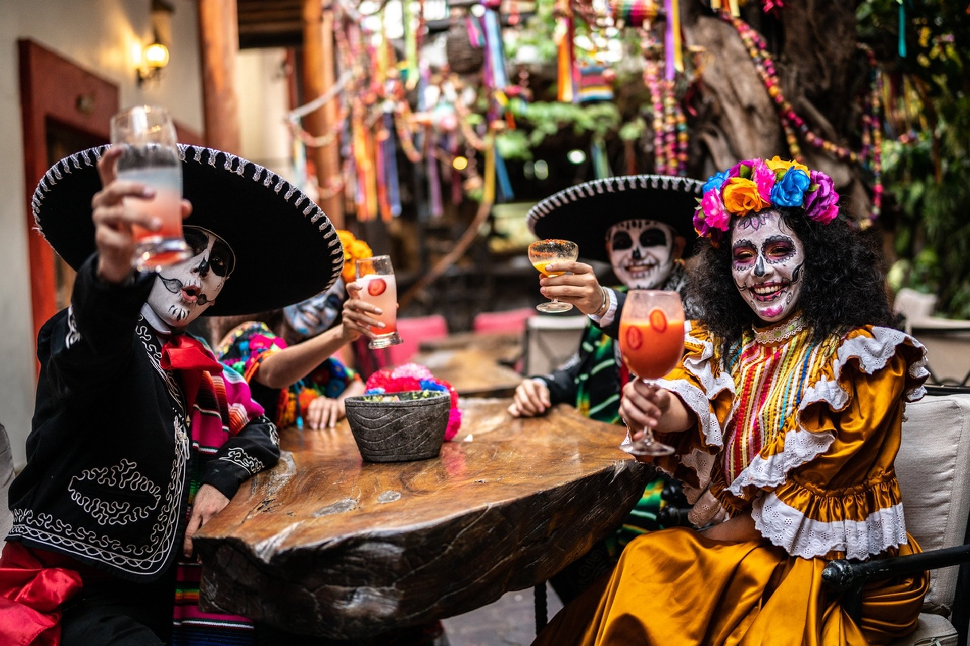 People in festive Day of the Dead attire, with face paint and colorful costumes, raise drinks around a wooden table, celebrating joyfully.