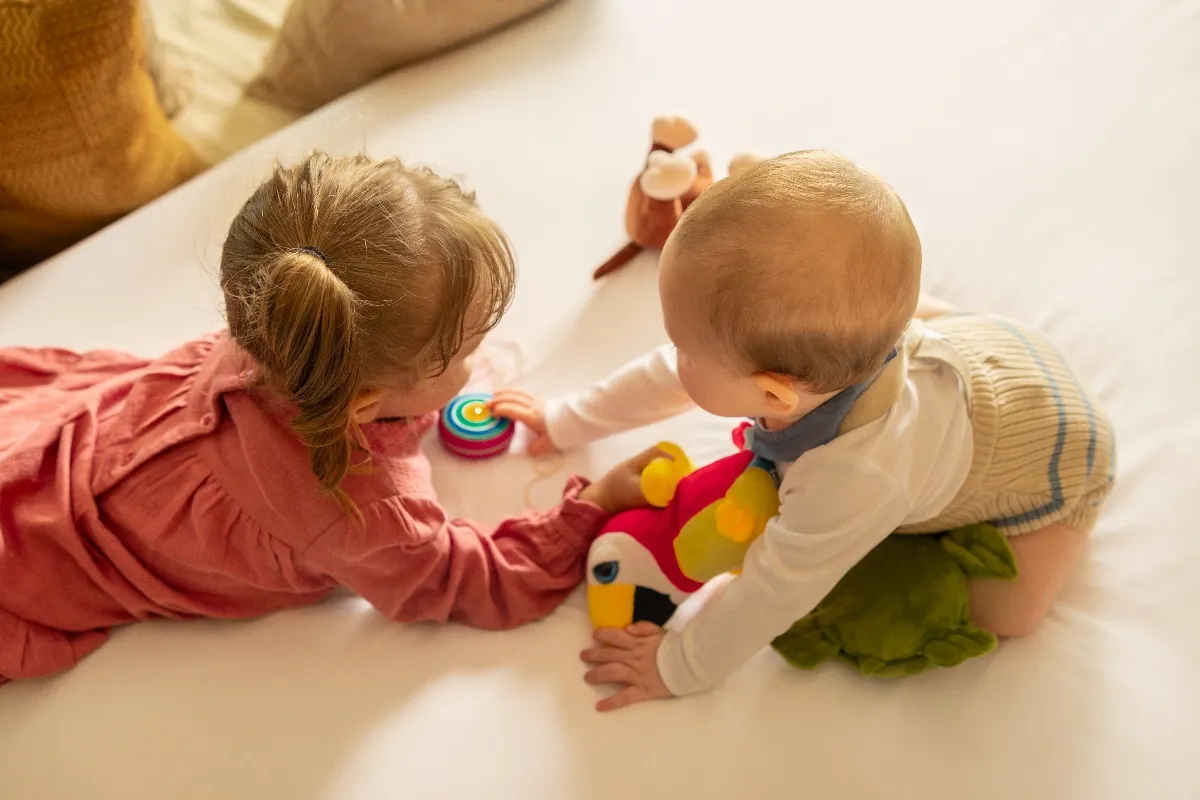 Two young children lying on a bed, playing with a colorful toy and stuffed animal, surrounded by soft light and warm colors.