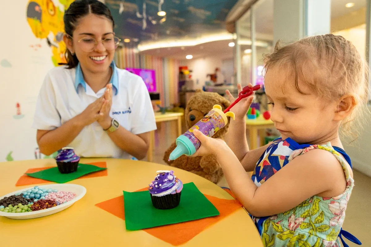 A young girl decorates cupcakes with sprinkles, assisted by a smiling staff member in a colorful children's activity room.