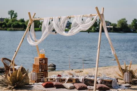 Beachfront picnic setup with draped canopy, cushions, lanterns, and a dining spread by the water.