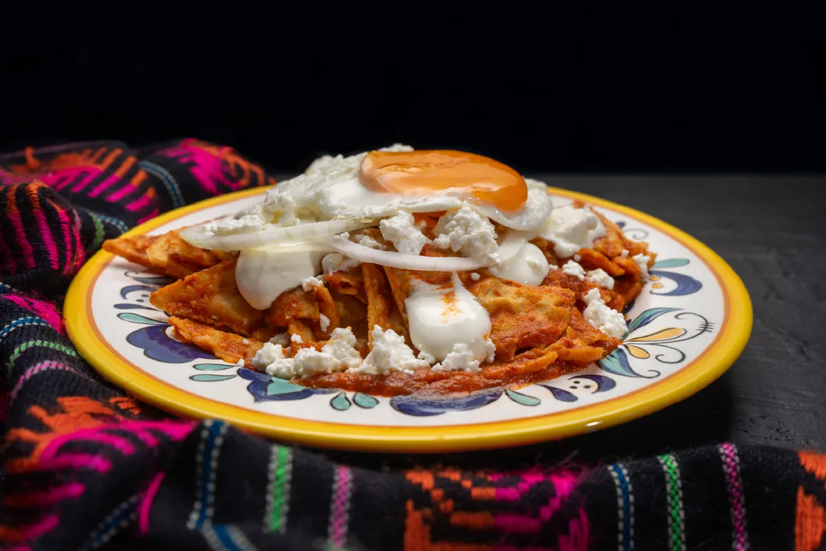 Plate of chilaquiles topped with a fried egg, cream, cheese, and onions, served on a colorful tablecloth.