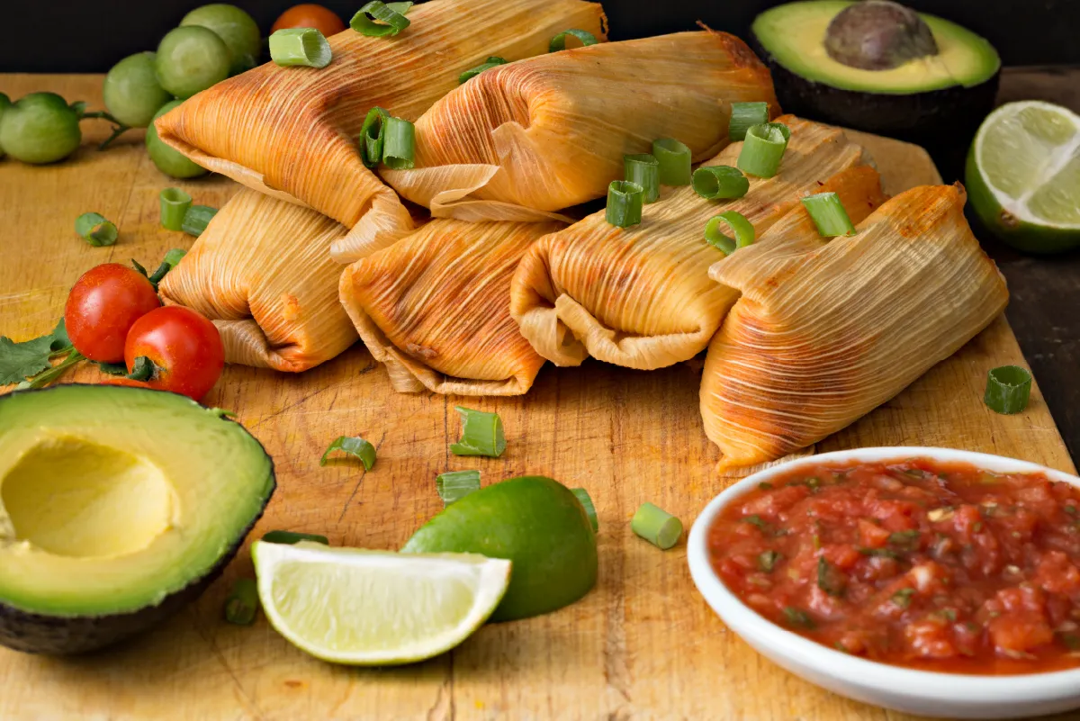 Tamales on a wooden board garnished with green onions, surrounded by avocados, limes, cherry tomatoes, and a bowl of salsa.