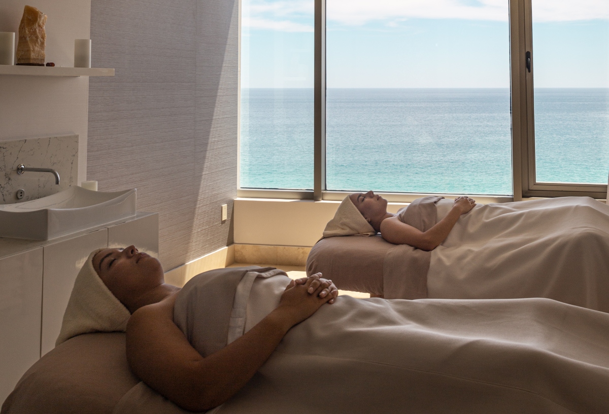 Two girls enjoying a relaxing spa treatment on massage tables, overlooking a serene ocean view through large windows.