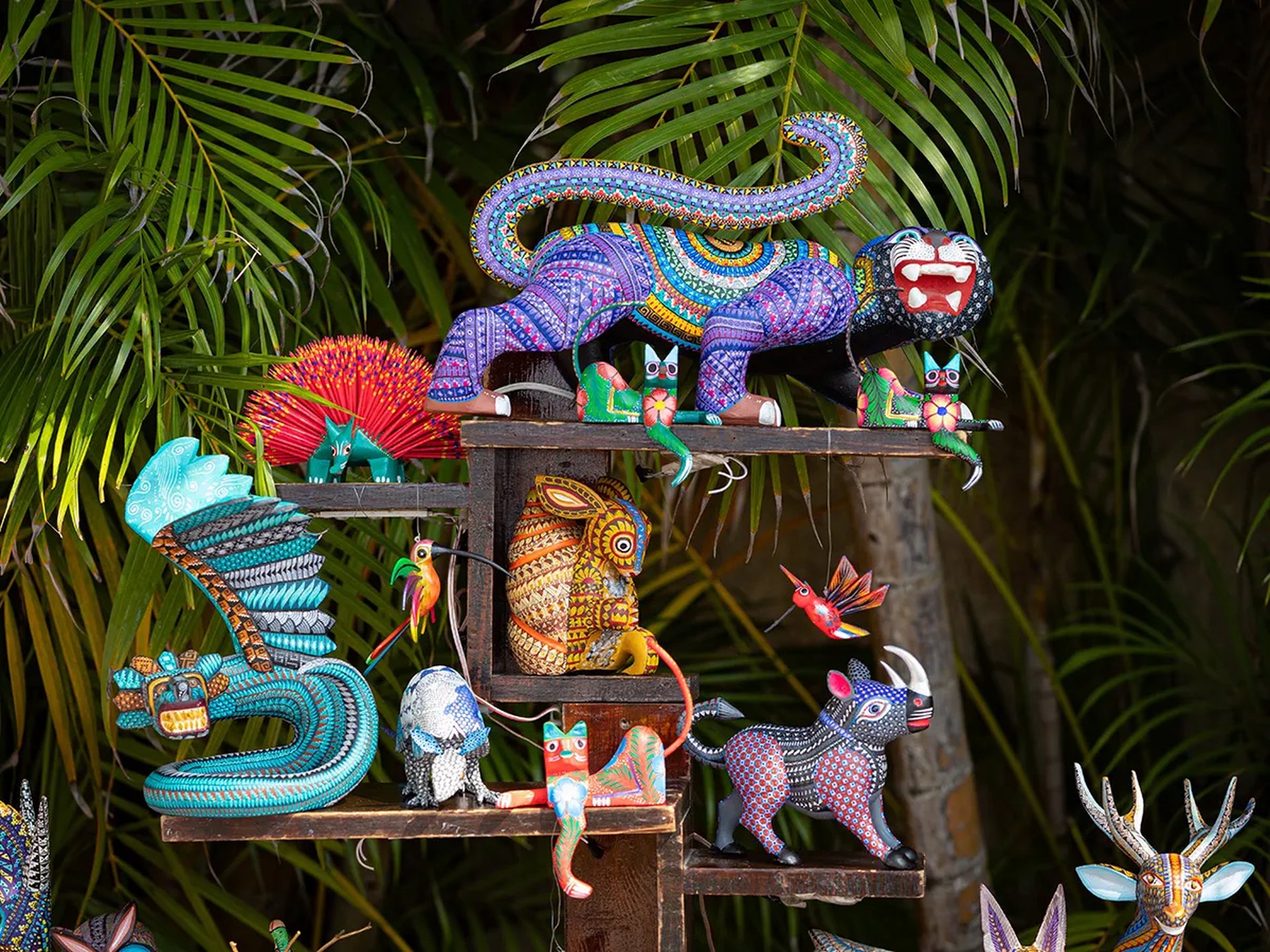 Colorful Mexican alebrijes, including a serpent, jaguar, and owl, displayed on wooden shelves with lush green foliage in the background.