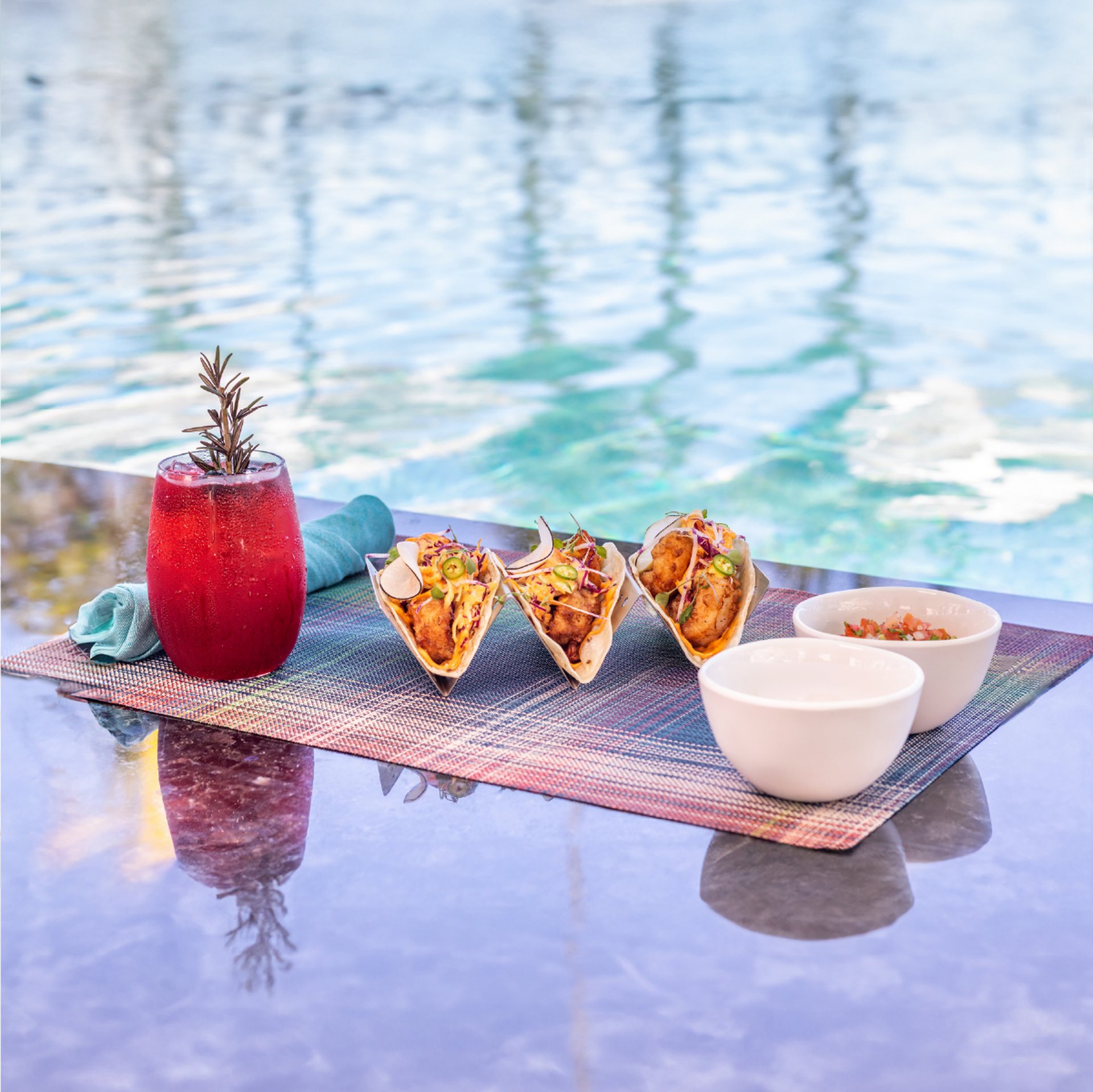 Poolside feast: a red rosemary cocktail with taco trio and bowls of salsa on a woven mat beside a turquoise pool.