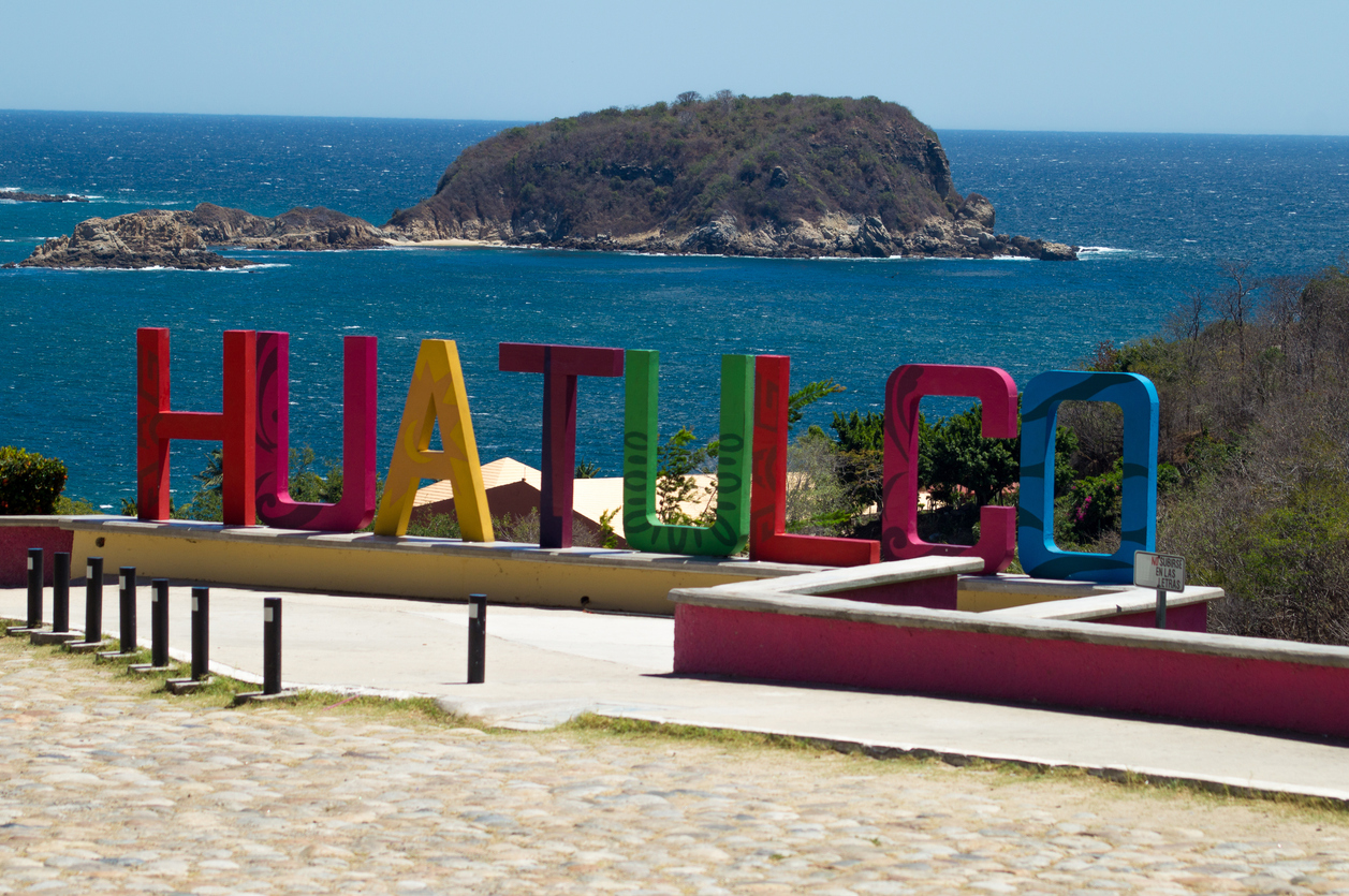 Colorful "Huatulco" sign overlooking the ocean, with a small island in the background under a clear blue sky.