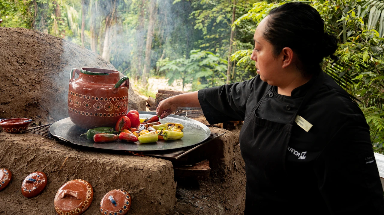 Chef cooking vibrant peppers and vegetables on an outdoor clay stove, surrounded by lush greenery.