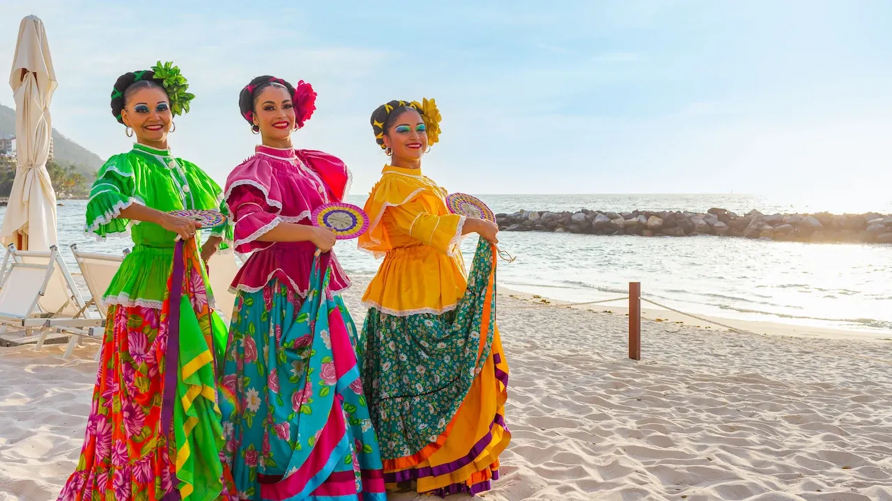 Three women in vibrant traditional Mexican dresses performing a dance on a sandy beach, with the ocean and sky in the background.