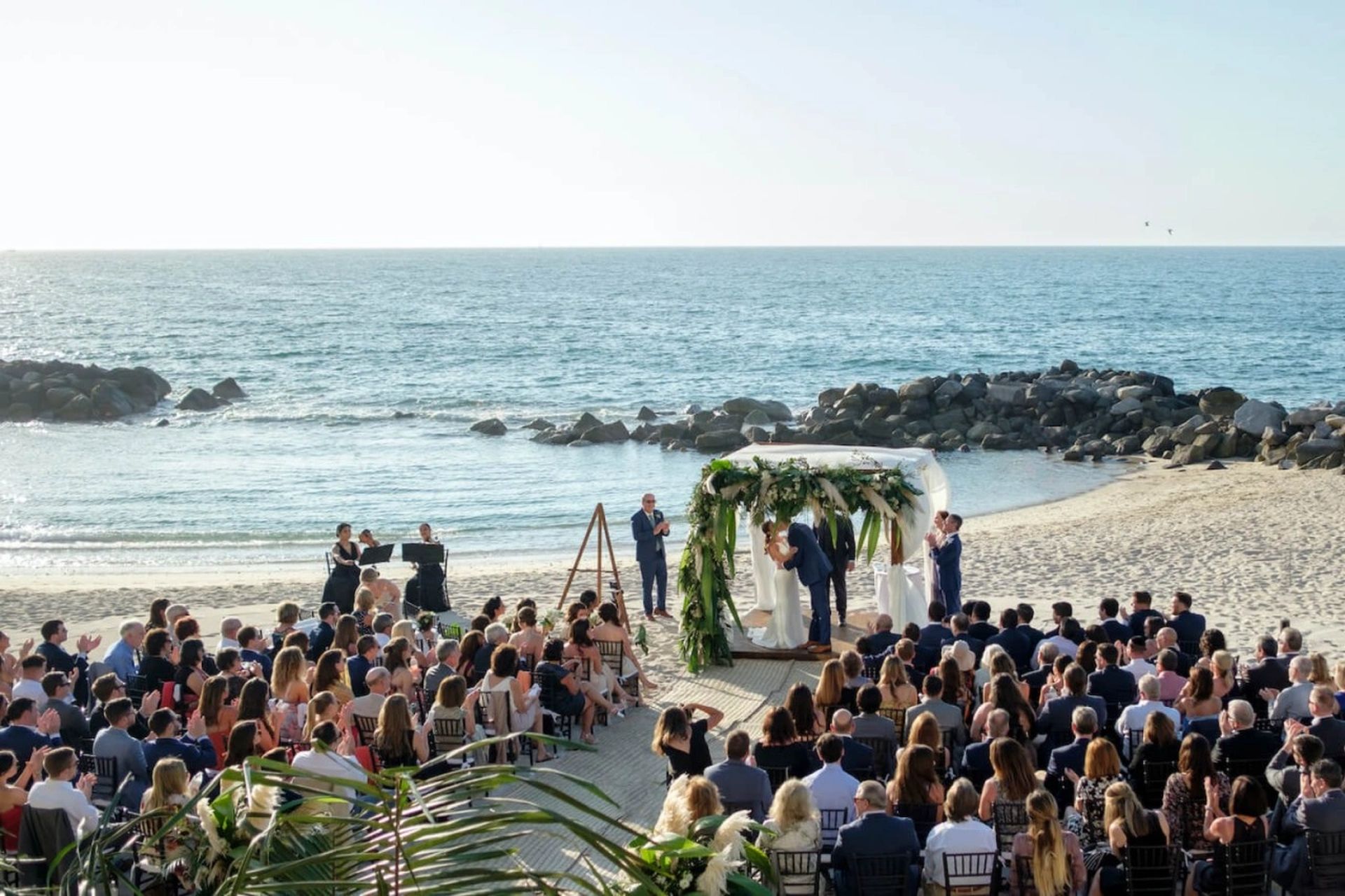 Beachfront wedding ceremony with guests seated, overlooking the ocean. A floral arch frames the couple, under a clear blue sky.
