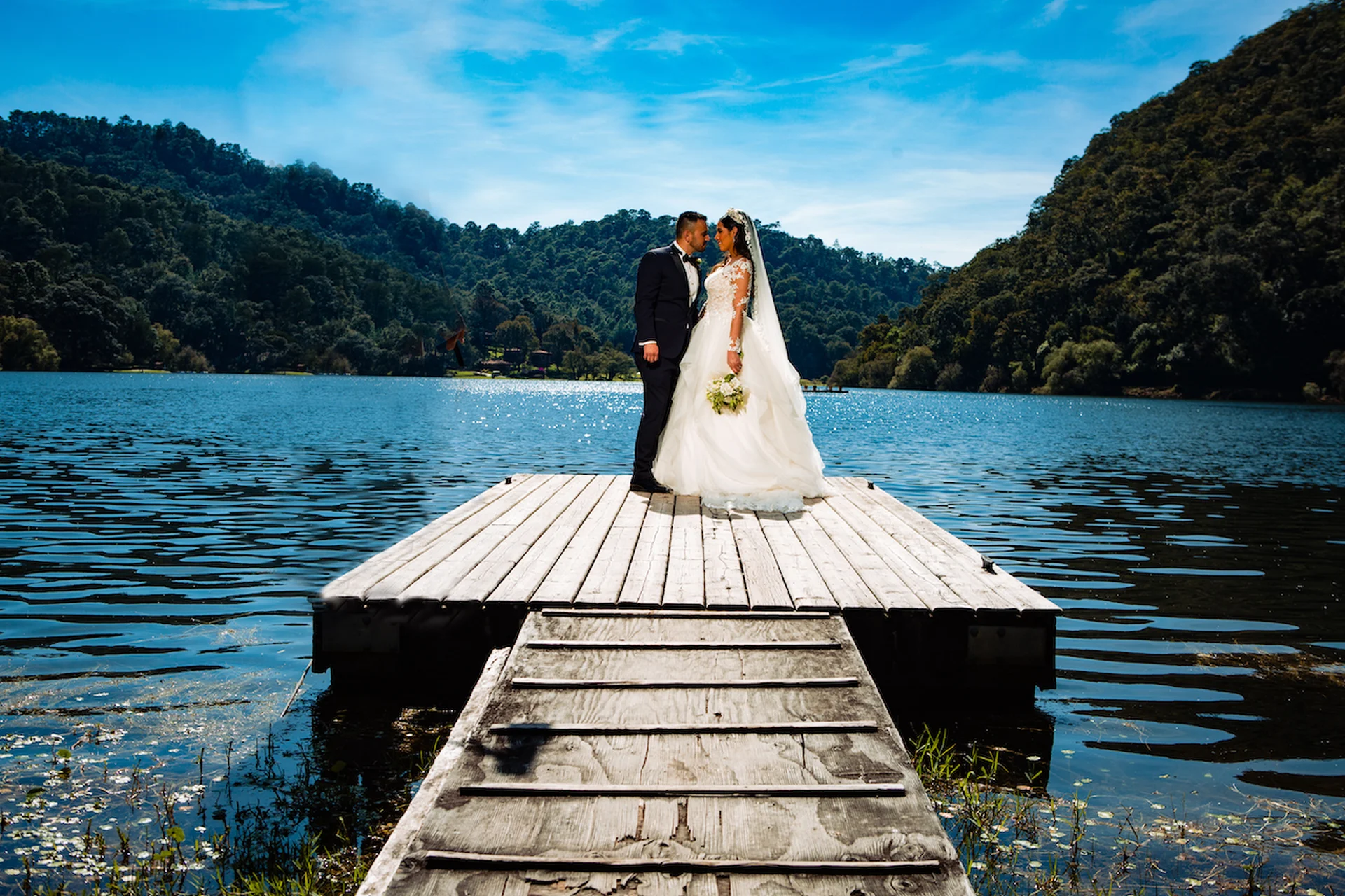 Bride and groom embracing at the end of a wooden dock on the lake, with forested mountains behind