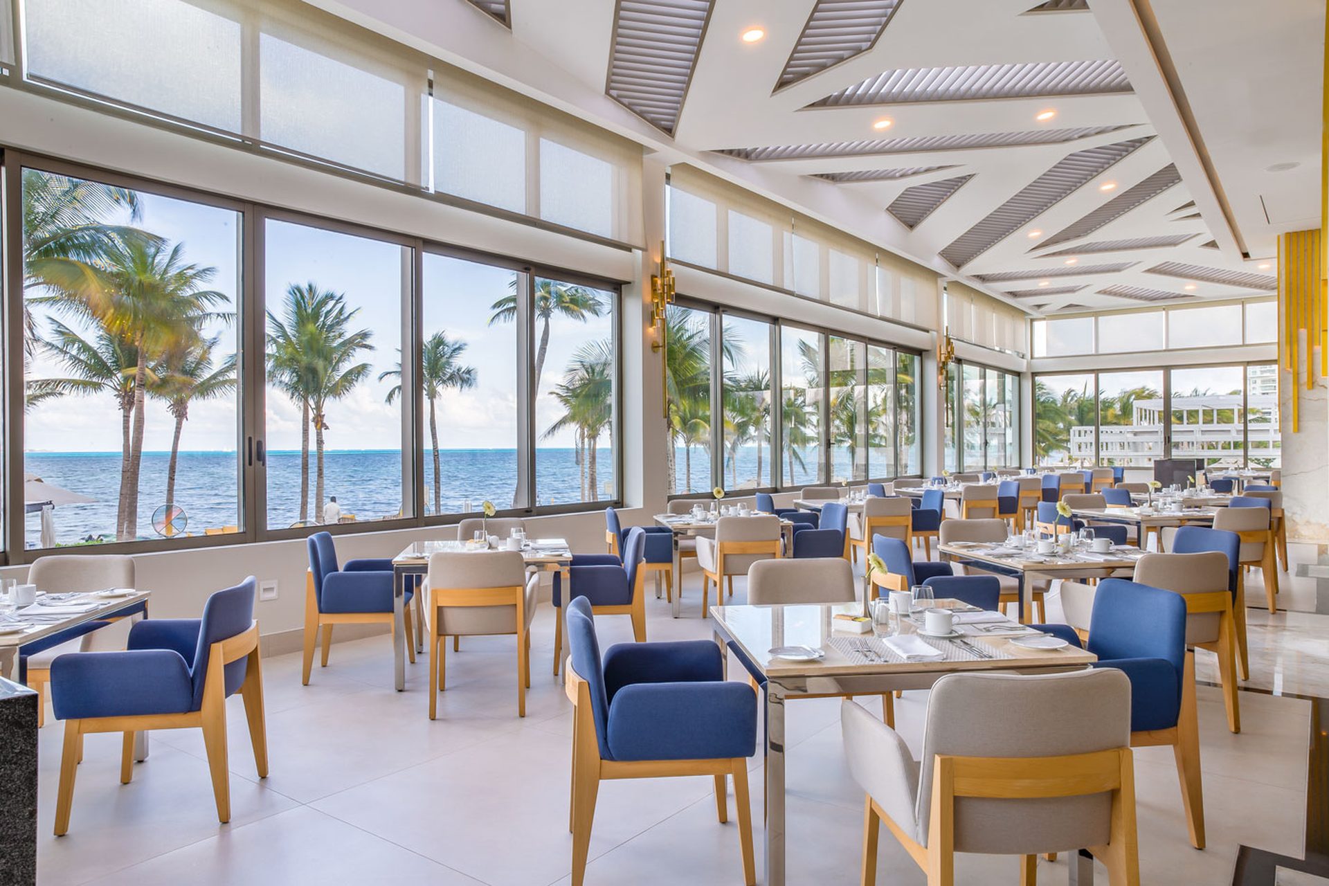 Elegant beachfront restaurant with panoramic ocean views, modern decor, blue and beige chairs, set tables, and palm trees outside.