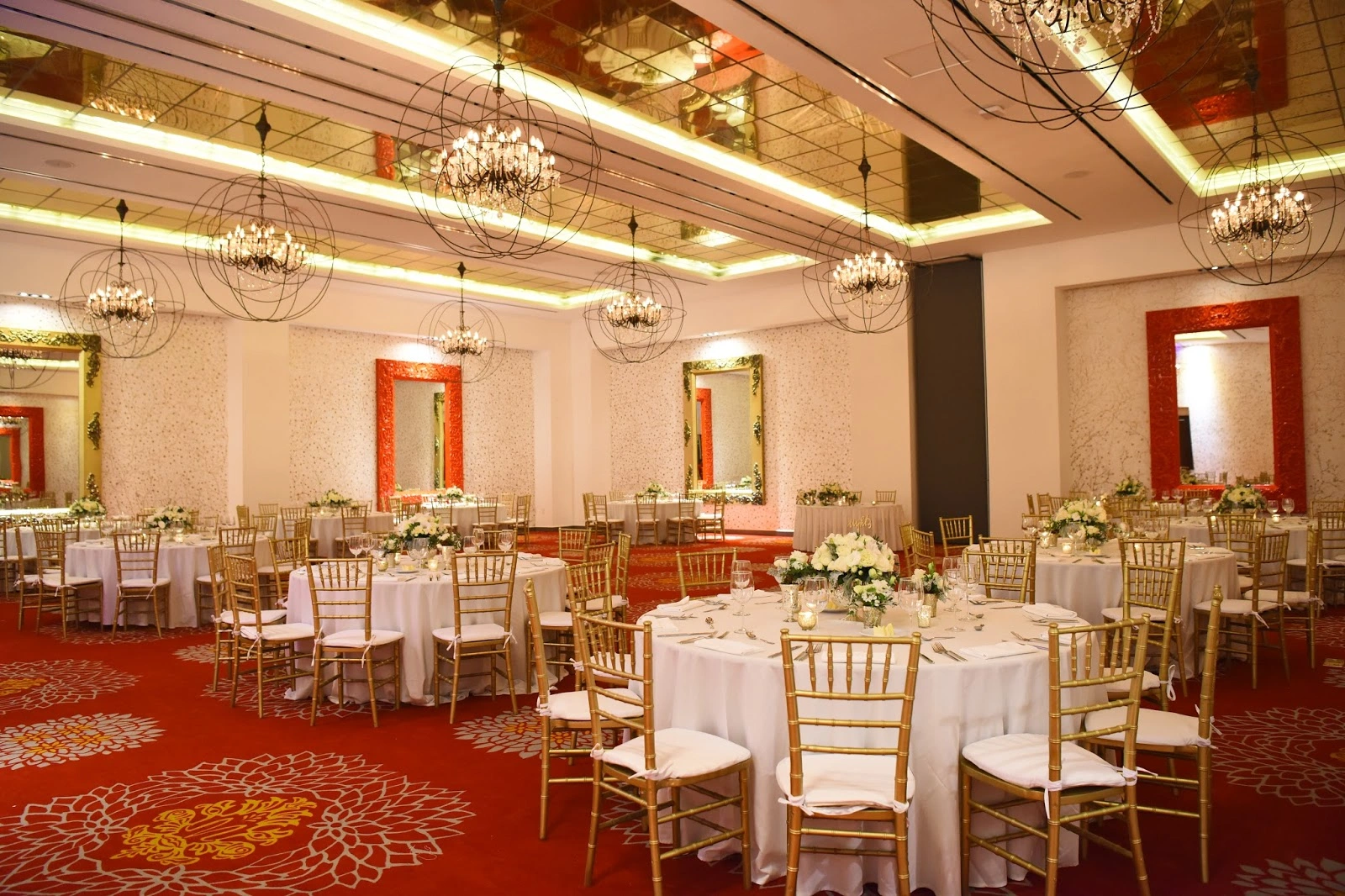 Elegant ballroom setup with round tables, white tablecloths, gold chairs, floral centerpieces, and ornate chandeliers reflecting in mirrors.