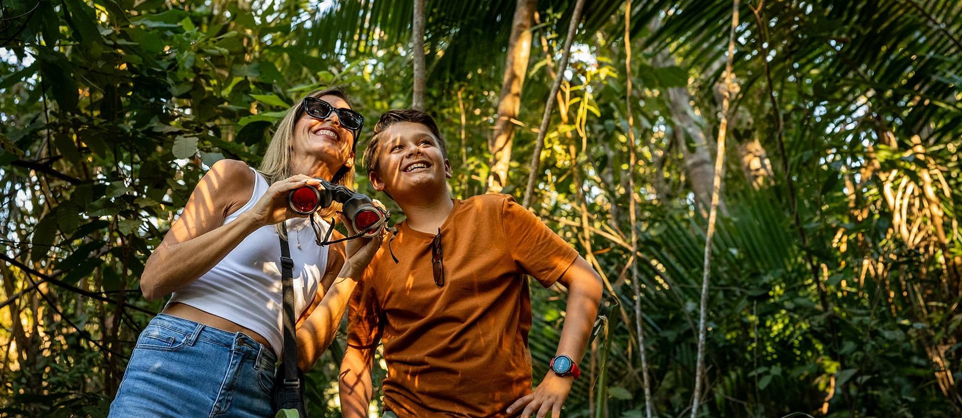 Woman and boy smiling in a lush forest, holding binoculars, surrounded by green foliage and dappled sunlight.